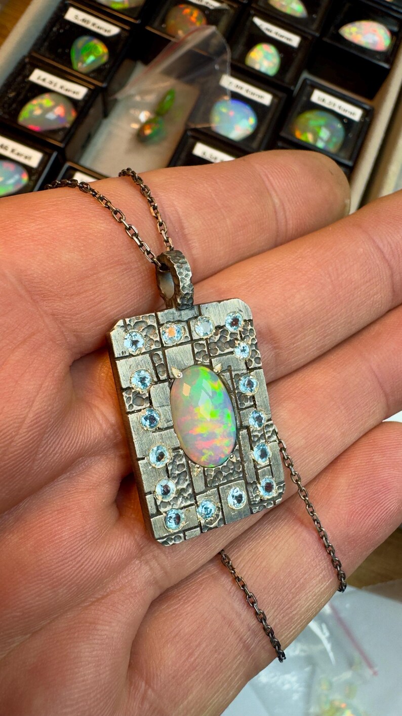 May include: A silver pendant necklace featuring a rectangular pendant with an oval opal gemstone at the center. The pendant is adorned with small, round, light blue gemstones and a textured design. The necklace chain is also silver.