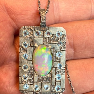 May include: A silver pendant necklace featuring a rectangular pendant with an oval opal center stone. The pendant is adorned with small, round, light blue gemstones and a textured border. The opal displays a play of colors.