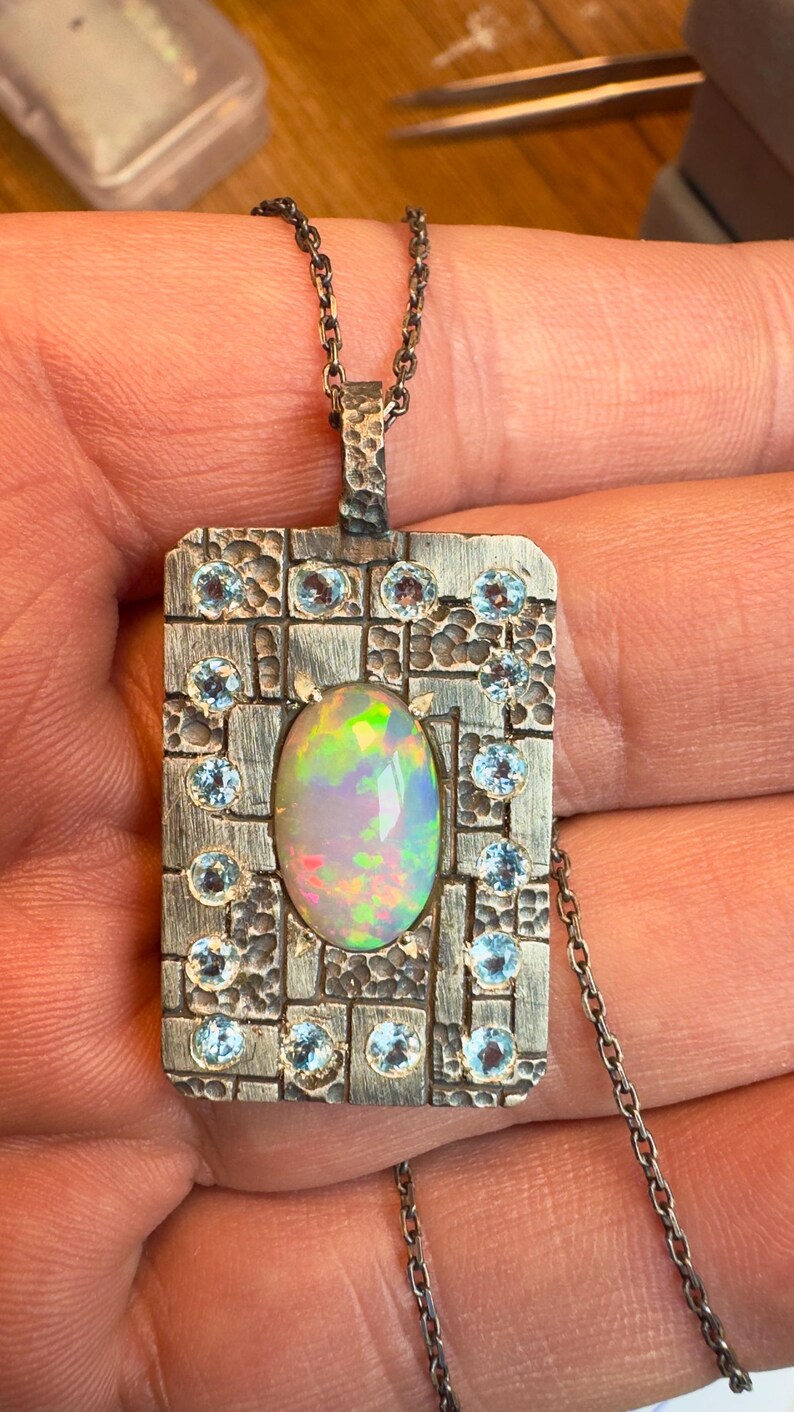 May include: A silver pendant necklace featuring a rectangular pendant with an oval opal center stone. The pendant is adorned with small, round, light blue gemstones and a textured border. The opal displays a play of colors.