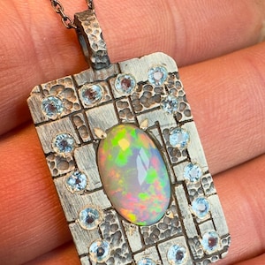 May include: A silver pendant necklace featuring a rectangular pendant with a textured surface. The pendant is adorned with a large, oval opal gemstone at the center, surrounded by smaller, round, light blue gemstones. The opal displays a vibrant play of colors.