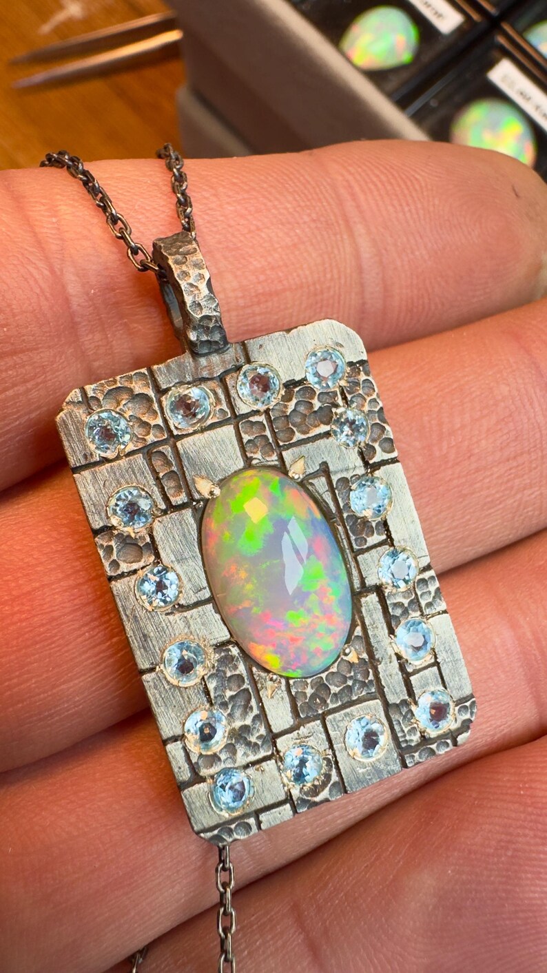 May include: A silver pendant necklace featuring a rectangular pendant with a textured surface. The pendant is adorned with a large, oval opal gemstone at the center, surrounded by smaller, round, light blue gemstones. The opal displays a vibrant play of colors.