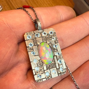 May include: A silver pendant necklace featuring an oval opal gemstone with iridescent colors. The rectangular pendant is adorned with small, round, light blue gemstones and a textured surface. The necklace has a delicate chain.