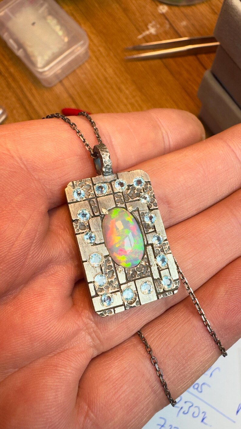 May include: A silver pendant necklace featuring an oval opal gemstone with iridescent colors. The rectangular pendant is adorned with small, round, light blue gemstones and a textured surface. The necklace has a delicate chain.