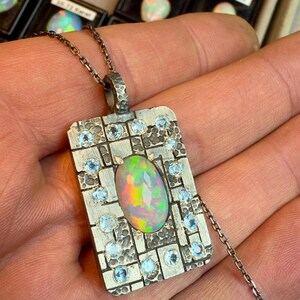 May include: A silver pendant necklace with a rectangular pendant featuring an oval opal gemstone. The pendant is adorned with small, round, light blue gemstones and hangs from a delicate chain. The opal displays a vibrant array of colors.