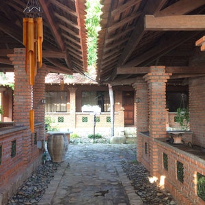 May include: A brick walkway with a stone path leading to a brick building with a wooden roof. A brown clay pot sits on the left side of the path. A wooden wind chime hangs from the roof.