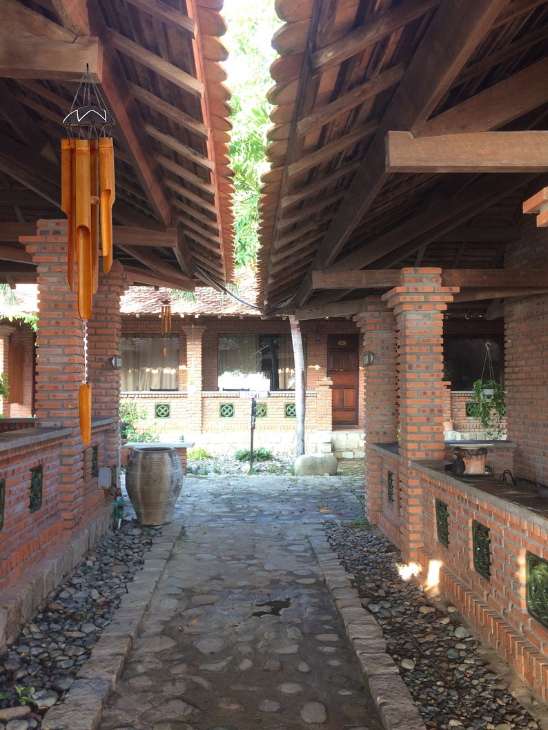 May include: A brick walkway with a stone path leading to a brick building with a wooden roof. A brown clay pot sits on the left side of the path. A wooden wind chime hangs from the roof.