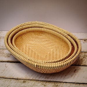 May include: Three woven oval baskets made of natural materials, stacked on top of each other. The baskets are light brown in color and have a natural, rustic look.