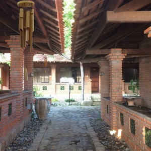 Puede incluir: Un camino de ladrillos con un sendero de piedra que conduce a una estructura de madera con techo de paja. Un carill&oacute;n de madera cuelga del techo de la estructura. El camino est&aacute; bordeado de peque&ntilde;as piedras.