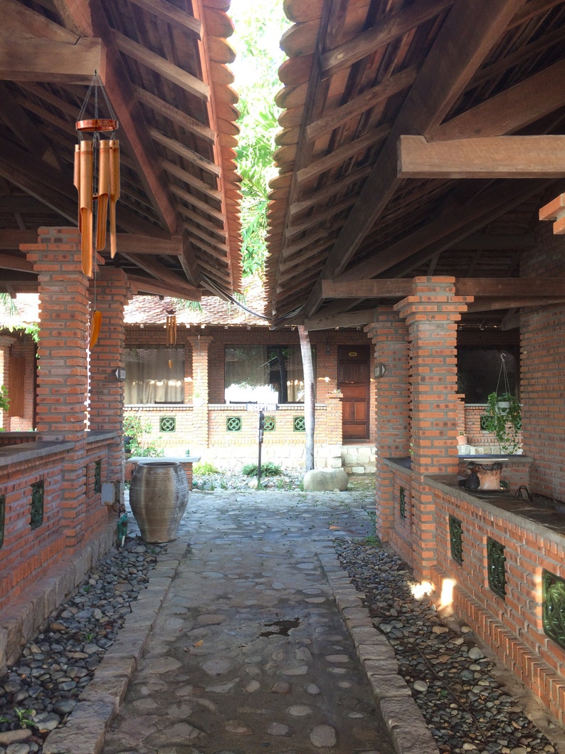 May include: A brick walkway with a stone path leading to a wooden structure with a brick wall on either side. The walkway is lined with small stones and there is a large clay pot on the left side of the path. The wooden structure has a wooden roof with exposed beams.