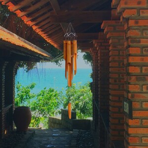 May include: A wooden wind chime hangs from the ceiling of a brick building with a view of the ocean and trees in the background.