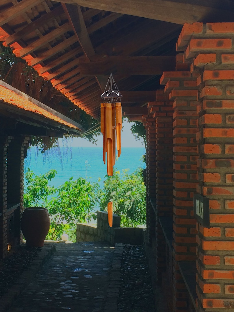 May include: A wooden wind chime hangs from the ceiling of a brick building with a view of the ocean and trees in the background.