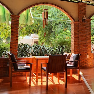 May include: A patio setting with three wooden chairs and a square wooden table. The chairs have brown cushions and the table is empty. The patio is covered by a brick archway and there is a wind chime hanging from the ceiling.