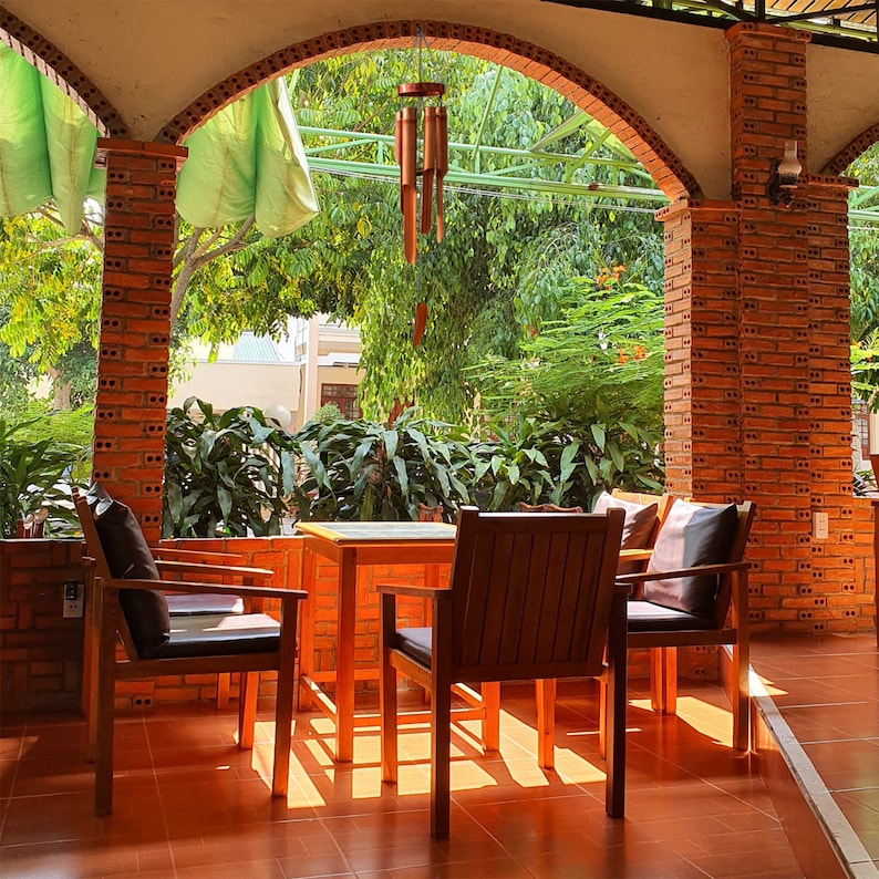 May include: A patio setting with three wooden chairs and a square wooden table. The chairs have brown cushions and the table is empty. The patio is covered by a brick archway and there is a wind chime hanging from the ceiling.