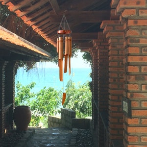 May include: A wooden wind chime hangs from the ceiling of a brick building. The chime is made of multiple wooden tubes and has a copper top. The chime is hanging in front of a doorway that leads to a view of the ocean.