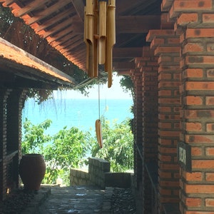 Puede incluir: Un carill&oacute;n de viento de madera con tubos de bamb&uacute; colgando de un marco de madera. El carill&oacute;n est&aacute; colgado en una puerta con vista al oc&eacute;ano y una pared de ladrillo.