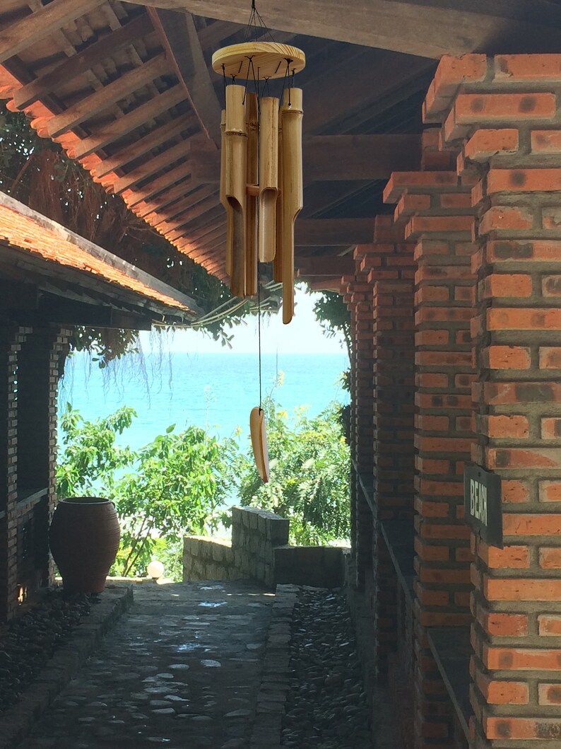 Puede incluir: Un carill&oacute;n de viento de madera con tubos de bamb&uacute; colgando de un marco de madera. El carill&oacute;n est&aacute; colgado en una puerta con vista al oc&eacute;ano y una pared de ladrillo.
