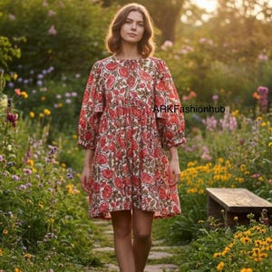 May include: A woman in a red and white floral print dress with three-quarter sleeves walks along a stone path in a garden. The dress has a round neck and a tiered skirt. The garden is full of colourful flowers and greenery.