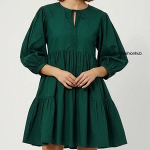 May include: A dark green dress with a round neckline and a keyhole detail. The dress features long, voluminous sleeves and a tiered skirt. The fabric appears to be a lightweight cotton or similar material. The dress has a relaxed, flowing silhouette.