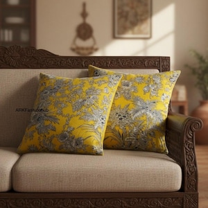 Puede incluir: Dos cojines decorativos sobre un sofá beige. Los cojines tienen un fondo amarillo brillante con un estampado floral gris y blanco. El sofá tiene una estructura de madera marrón oscuro con detalles tallados.