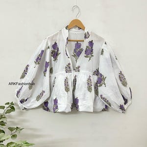 Può includere: Blusa bianca in cotone con stampa floreale viola e vita arricciata. La blusa ha una scollatura a V e maniche lunghe.