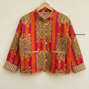 Puede incluir: Una chaqueta acolchada con un vibrante patrón de rayas verticales naranjas y magenta con un estampado de tigre repetitivo. La chaqueta tiene cuello mao, mangas largas con puños a rayas, cinco botones claros y dos bolsillos delanteros. El texto ARKFashionhub es visible en el lado derecho.
