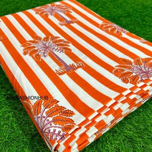 May include: Folded fabric with a repeating pattern of orange and white stripes and palm tree illustrations. The palm trees are in shades of orange and purple. The fabric is folded neatly, showing the pattern on the top layer.