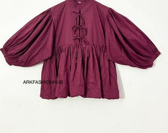 May include: A burgundy blouse with a high, ruffled collar and voluminous sleeves. The blouse features a tie-up detail at the neckline and a gathered waist. The text "ARKFASHIONHUB" is visible at the bottom of the image. The blouse is hanging on a wooden hanger.