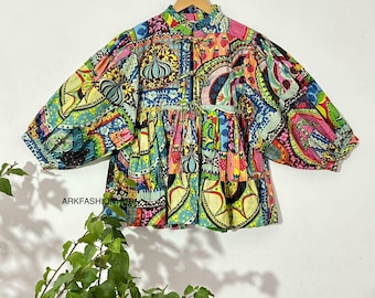 May include: A colorful, patterned top with a high collar and three-quarter sleeves. The top features a patchwork design with various shapes and colors, including pink, blue, yellow, and green. The garment is hanging on a wooden hanger.