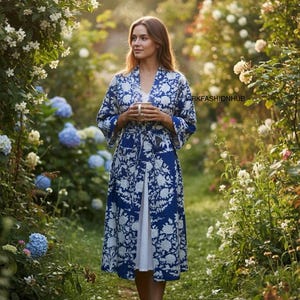 May include: A woman wearing a blue and white floral print robe, holding a cup, stands in a garden. The robe has a matching belt and is over a white dress. The garden features lush greenery and blooming flowers.