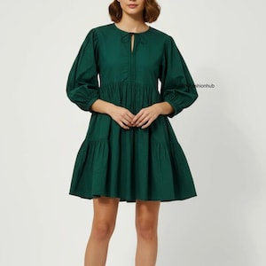 May include: A dark green mini dress with a keyhole neckline and long, puffed sleeves. The dress has a tiered skirt and a loose fit. The fabric looks like a lightweight cotton. The dress is a solid, deep green colour.