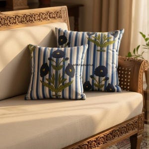 Può includere: Due cuscini quadrati con strisce verticali blu e bianche e motivi floreali. I cuscini presentano un fiore blu scuro con foglie verdi. I cuscini sono su un divano beige con una cornice in legno intagliato.