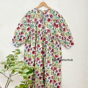 Può includere: Abito bianco a maniche lunghe con un vivace motivo floreale in rosso, blu e verde. L'abito presenta uno scollo tondo con fiocco, vita arricciata e maniche a tre quarti. Il testo "ARKfashionhub" è visibile.