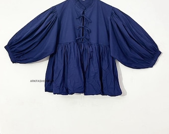 May include: A navy blue blouse with a high collar and long, voluminous sleeves. The blouse features a series of fabric ties down the front and a gathered, flared hem. The garment is hanging on a wooden hanger against a white background.