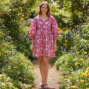 Peut inclure: Une femme porte une robe courte rose à motifs floraux blancs et manches bouffantes. La robe a un col en V et arrive au genou. Elle porte des sandales et marche sur un sentier bordé de fleurs et de verdure.