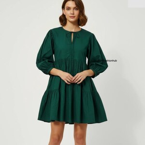 May include: A dark green dress with a round neckline and long, puffed sleeves. The dress has a keyhole detail at the chest and a tiered skirt. The dress is made of a solid-coloured fabric and has a casual, yet stylish design.