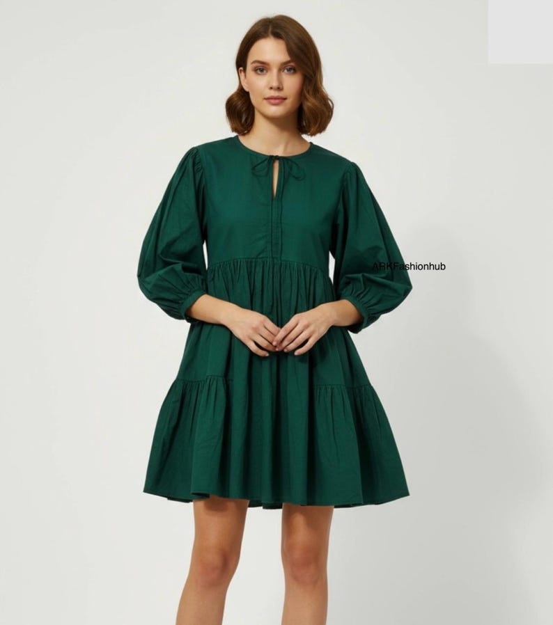 May include: A dark green dress with a round neckline and long, puffed sleeves. The dress has a keyhole detail at the chest and a tiered skirt. The dress is made of a solid-coloured fabric and has a casual, yet stylish design.
