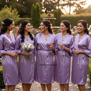 May include: Five women wearing matching lavender satin robes, standing outdoors in a garden setting. The robes have a wrap-around design with a tie closure. One woman holds a bouquet of white flowers.