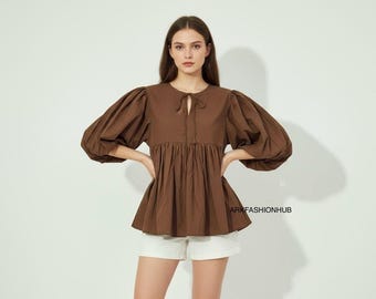 May include: A brown blouse with voluminous, elbow-length sleeves and a tie detail at the neckline. The blouse has a gathered waist and is paired with white shorts. The text "ARKFASHIONHUB" is visible on the lower right.