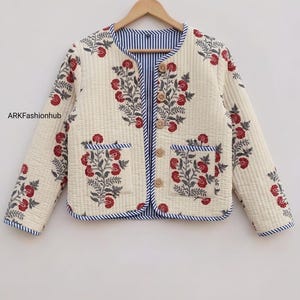 Puede incluir: Una chaqueta acolchada en color crema con estampado floral rojo y gris, con ribete a rayas azules y blancas, mangas largas, dos bolsillos delanteros y cinco botones de madera clara. El texto ARKFashionhub es visible en el lado izquierdo.