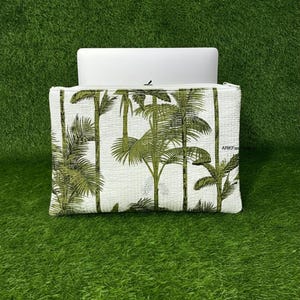 Pode incluir: Capa branca para laptop com um padrão de palmeiras verdes. A capa retangular tem um fecho de correr. O fundo é uma superfície verde e gramada.