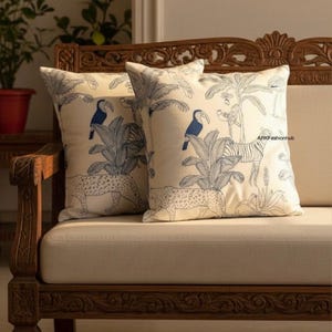 May include: Two square cushions with a cream background and a blue jungle design. The design features toucans, leopards, palm trees, and zebras. The cushions are on a wooden sofa with carved details.