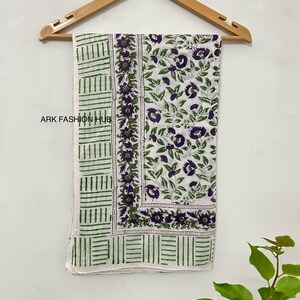 May include: A white textile with a floral pattern and green geometric border is displayed on a wooden hanger. The floral design features purple flowers and green leaves. The text "ARK FASHION HUB" is visible.