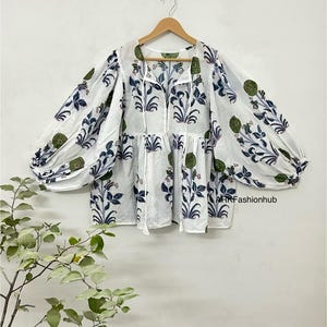 Könnte beinhalten: Weiße Bluse mit blauem und grünem Blumenmuster. Das langärmlige Oberteil hat einen runden Ausschnitt und eine geraffte Taille. Die Ärmel sind ballonartig und am Handgelenk gerafft. Die Bluse hängt an einem Holzkleiderbügel.