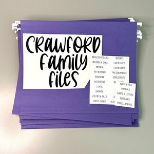 Puede incluir: Una pila de carpetas de archivos moradas con pesta&ntilde;as blancas etiquetadas con categor&iacute;as para organizar documentos familiares. La carpeta delantera tiene una etiqueta blanca con el texto "Crawford Family Files" en negro.