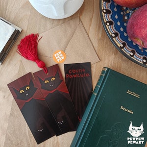Könnte beinhalten: Ein Stillleben mit einem grünen Buch mit dem Titel "Dracula", zwei schwarzen Lesezeichen mit Katzenmotiven und einem Lesezeichen mit der Aufschrift "Count Pawcula". Ein roter Quastenanhänger und ein orangefarbener Aufkleber sind ebenfalls zu sehen.