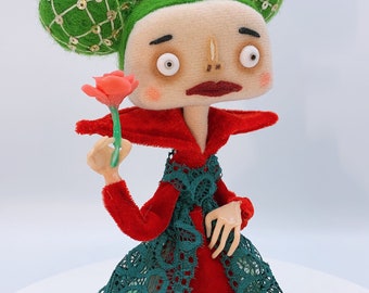 Puede incluir: Una muñeca de fieltro hecha a mano con cabello verde y un vestido de terciopelo rojo. La muñeca sostiene una flor rosa y tiene una expresión caprichosa.