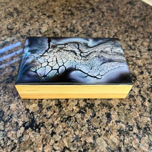 May include: A rectangular wooden box with a lid displaying an abstract design in black, white, and blue hues. The box has a natural wood base and a glossy finish on the lid. The design resembles a fluid art pattern.