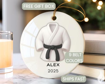 May include: A round white ceramic ornament featuring a 3D-style graphic of a white martial arts uniform with a black belt. The text ALEX and 2025 are printed in black below the uniform. A gold string is attached to the top for hanging purposes.