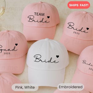 May include: A collection of pink and white baseball caps. The pink caps have the words "Team Bride", "Bride 2025", "Squad 2025", and "Bridesmaid 2025" embroidered on them. One white cap has "Bride" embroidered on it. All caps have a black heart.