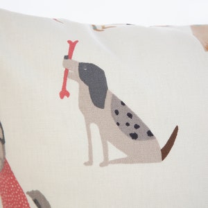 K&ouml;nnte beinhalten: Ein wei&szlig;er Kissenbezug mit einem sich wiederholenden Muster aus Hunden. Die Hunde sind im Cartoon-Stil illustriert und sind verschiedener Rassen. Die Hunde sind in Braun-, Schwarz- und Wei&szlig;t&ouml;nen gehalten. Ein Hund h&auml;lt einen roten Knochen im Maul.